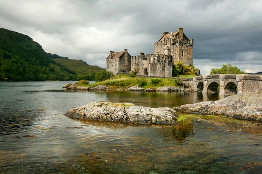 Sun shines through clouds on Eilean Donan castle with rocks in foreground loch. Dornie, Scotland