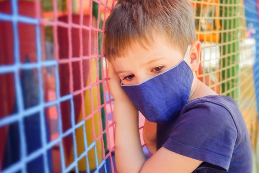 Kid in a medical mask standing near a closed playground. Child in face mask in front of playground. Bored boy in medical mask near playground during covid-19 global pandemic. Coronavirus outbreak