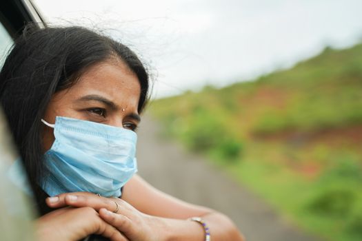 Sad Young girl in medical mask to protect from coronavirus, covid-19 or pollution and watching out from moving car window - concept of travel, vacation and road trip