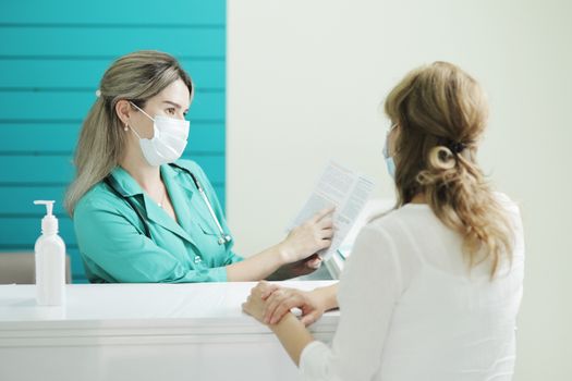 A female doctor or nurse wearing a medical mask talks to a female patient in the waiting room of a hospital. Disinfector on a rack