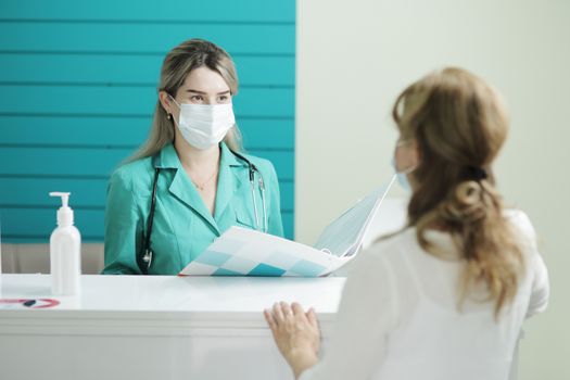 A female doctor or nurse wearing a medical mask talks to a female patient in the waiting room of a hospital. Disinfector on a rack