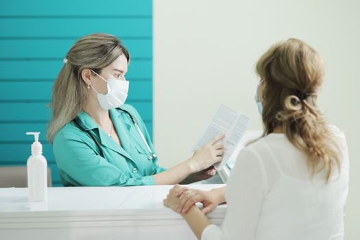 A female doctor or nurse wearing a medical mask talks to a female patient in the waiting room of a hospital. Disinfector on a rack