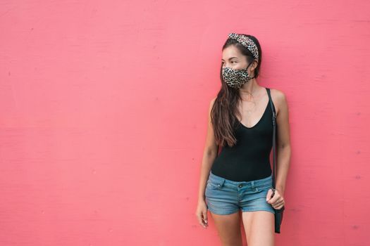 Mask wearing is mandatory in indoor spaces and public park for COVID 19. Asian woman is using face mask with fashion leopard fabric on pink wall background. City lifestyle outside for corona virus.