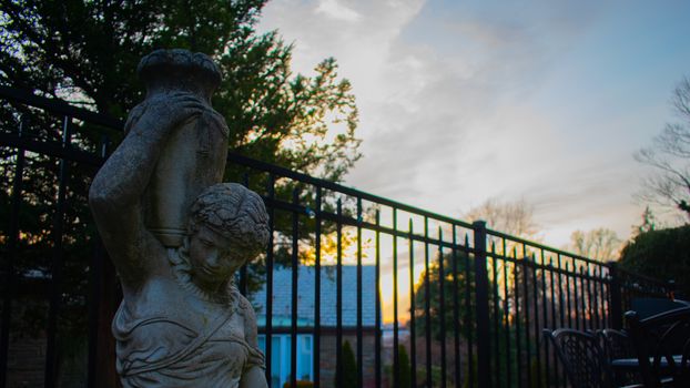 A Statue of a Woman Holding a Vase With a Fence and Sunset in the background