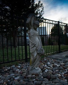 A Statue of a Woman Holding a Vase With a Fence and Sunset in the background