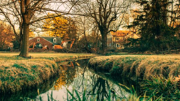 A Small Creek in a Field in a Rural Neighborhood With Houses and Heavy Machinery in the Background With the Scene Reflecting Off the Water