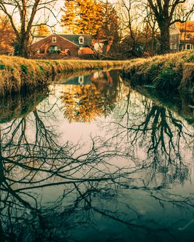 A Small Creek in a Field in a Rural Neighborhood With Houses and Heavy Machinery in the Background With the Scene Reflecting Off the Water