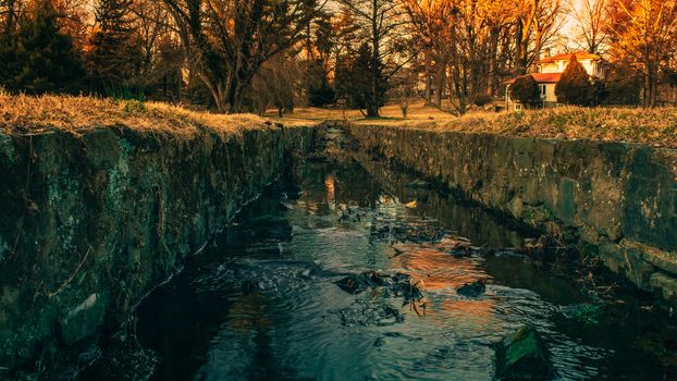 A Small Man-Made Creek With Cobblestone Walls on the Sides, trees and an Orange Sunset in the background.