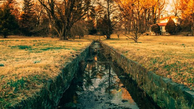 A Small Man-Made Creek With Cobblestone Walls on the Sides, trees and an Orange Sunset in the background.