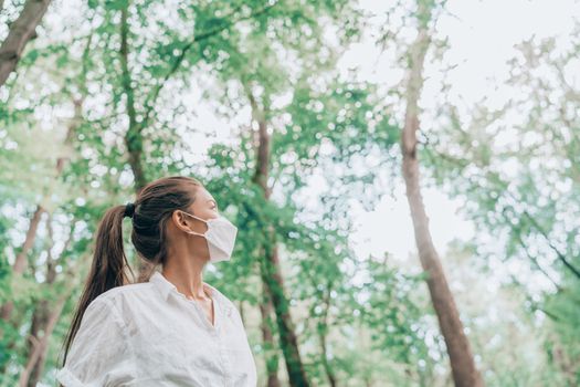 Woman hiking in forest nature wearing face mask while walking outdoors, looking up at trees in hope. Clean air, sustainability, eco-friendly masks for coronavirus protection concept.