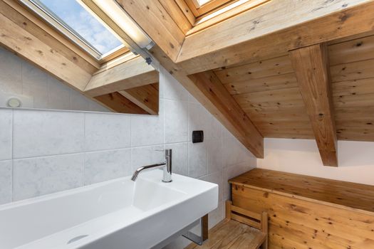 Practical and cozy bathroom witj wooden furnishings. Roof with exposed wooden beams and sky window. Italian style.