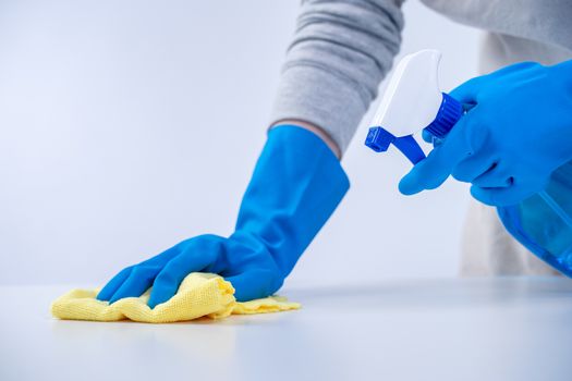 Young woman housekeeper in apron is cleaning, wiping down table surface with blue gloves, wet yellow rag, spraying bottle cleaner, closeup design concept.