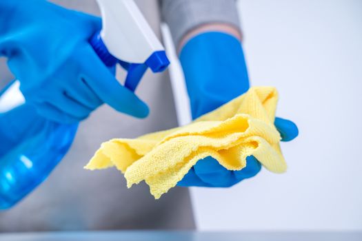 Young woman housekeeper in apron is cleaning, wiping down table surface with blue gloves, wet yellow rag, spraying bottle cleaner, closeup design concept.