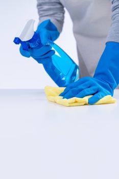 Young woman housekeeper in apron is cleaning, wiping down table surface with blue gloves, wet yellow rag, spraying bottle cleaner, closeup design concept.