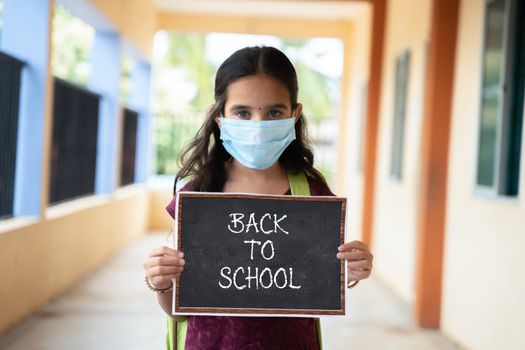 Young girl in medical mask holding back to school signage board at corridor - concept of school reopen, lifestyle and new normal