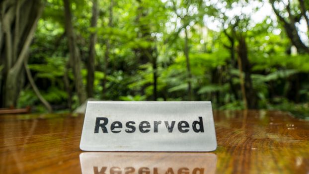 Reserved sign made out stainless steel plate on a laminated wooden table of a restaurant with trees and forest in the background.