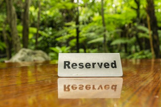 Reserved sign made out stainless steel plate on a laminated wooden table of a restaurant with trees and forest in the background.