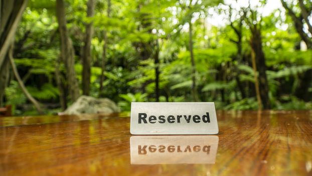 Reserved sign made out stainless steel plate on a laminated wooden table of a restaurant with trees and forest in the background.