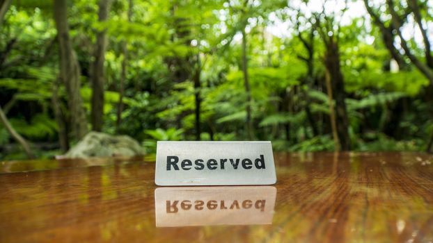 Reserved sign made out stainless steel plate on a laminated wooden table of a restaurant with trees and forest in the background.