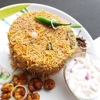 Colorful Delicious yummy prawn briyani with prawn fry in center with prawn fry and raita plated beautifully in white plate with bayleaf and it's one of favorite restaurant cuisine food