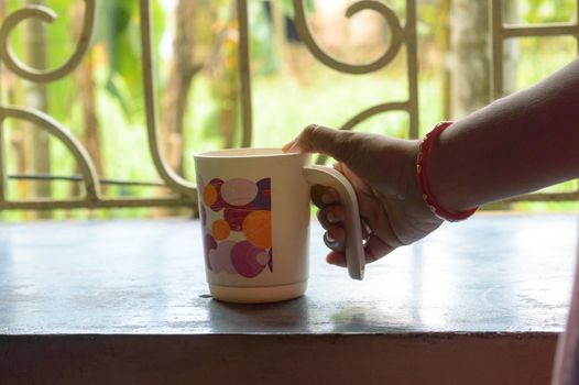 Woman Hand holding coffee cup. Close Up Of Human Hand Holding a Coffee Cup. Female hand hold take away coffee mug At Home. Morning sunny summer day in window sunlight background.