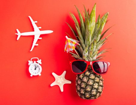 Summer Pineapple Day Concept; Top view flat lay of funny pineapple wear red sunglasses; model plane; starfish and clock alarm isolated on red background; Holiday summertime in tropical