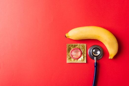 World sexual health or Aids day, Top view flat lay condom in wrapper pack, banana and doctor stethoscope, studio shot isolated on a red background, Safe sex and reproductive health concept