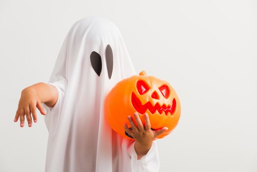 Funny Halloween Kid Concept, little cute child with white dressed costume halloween ghost scary he holding orange pumpkin ghost on hand, studio shot isolated on white background