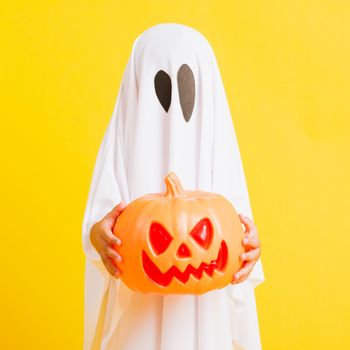 Funny Halloween Kid Concept, little cute child with white dressed costume halloween ghost scary he holding orange pumpkin ghost on hand, studio shot yellow on white background