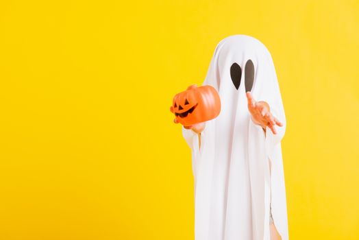 Funny Halloween Kid Concept, little cute child with white dressed costume halloween ghost scary he holding orange pumpkin ghost on hand, studio shot yellow on white background