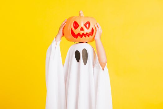 Funny Halloween Kid Concept, little cute child with white dressed costume halloween ghost scary he holding orange pumpkin ghost on hand, studio shot yellow on white background