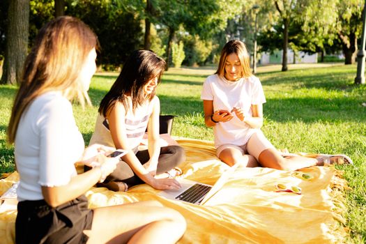 Three beautiful young female caucasian friends sitting on the grass at the park in the morning using smartphone - New technology habits of people - Using internet outdoor to smart working