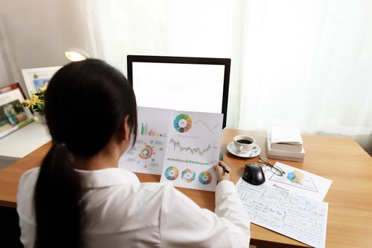 Rear view of businesswoman working in office with computer holding chart report paper and looking. businesswoman working at home with paper and empty PC screen. Business and finance concept.