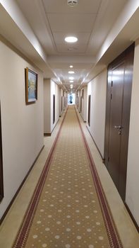 Modern hotel corridor with brown carpet Walls and ceiling white. Room numbers are painted on the wall next to the door. The lights in the ceiling are equipped with motion detectors for saving energy.