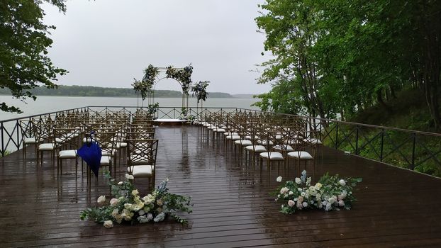 Wedding. Ceremony. Wedding arch and white chairs. Arch made from wood decorated with beige cotton textile, red, white and purple flowers and greenery, standing in ceremony area in woods.