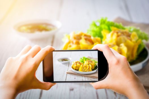Food photographer and social media concept. Hand holding black smartphone and take dumpling or dim sum photo for share on blog, website, and social media channel.