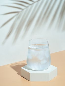 Cold water with ice in tumbler glass on hexagon pedestal. Mockup for drink in fashion trendy style with tropical leaf shadow. Modern still life with glass. Copy space. Health, beverage, summer concept