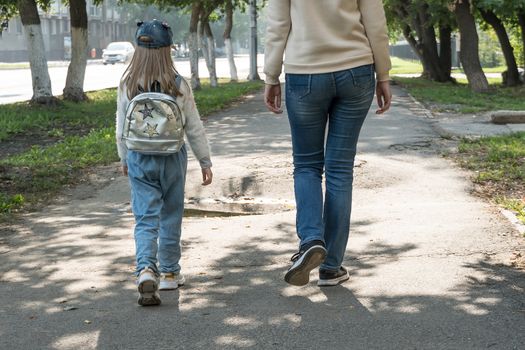 The mother takes the child's daughter to school. A girl with a backpack on her back is walking with her mother on the street.