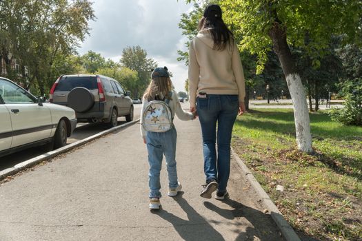 The mother takes the child's daughter to school. A girl with a backpack on her back is walking with her mother on the street.