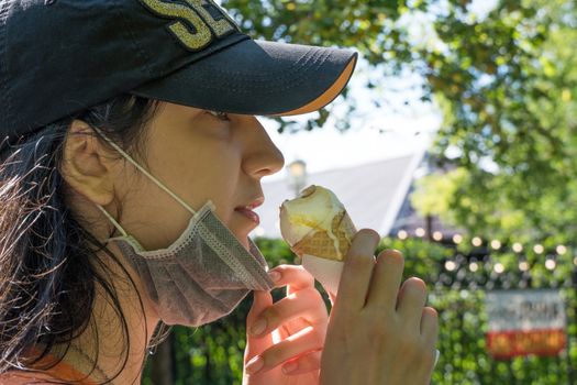 A young girl of Caucasian appearance eats an ice cream cone in a medical mask on the street during a quarantine for a coronavirus infection in summer. The weakening of the quarantine in the countries