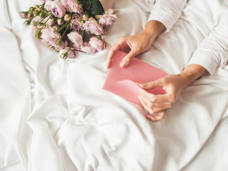 Woman is lying on crumpled bed sheet. She received bouquet of lilac colored roses and letter in pink envelope. Cute present for Valentine Day or Happy Birthday surprise.