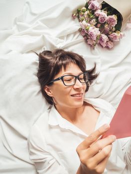 Young smiling woman is lying on crumpled bed sheet. She received bouquet of lilac colored roses and letter in pink envelope. Cute present for Valentine Day or Happy Birthday surprise.