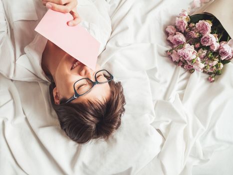 Young smiling woman is lying on crumpled bed sheet. She received bouquet of lilac colored roses and letter in pink envelope. Cute present for Valentine Day or Happy Birthday surprise.