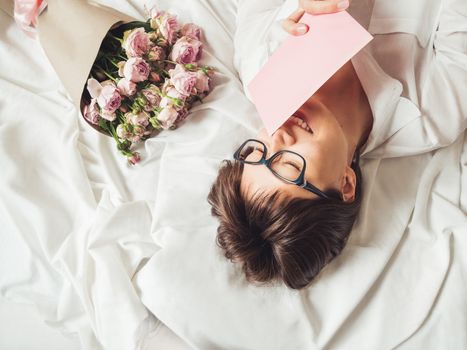 Young smiling woman is lying on crumpled bed sheet. She received bouquet of lilac colored roses and letter in pink envelope. Cute present for Valentine Day or Happy Birthday surprise.