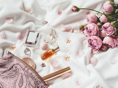 Cosmetic bag on crumpled white fabric background. Decorative cosmetic - perfume, mascara, eye and brow shadows. Cute and fashionable jewelry accessories. Pink roses and petals.