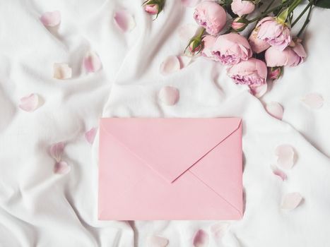 Roses and petals on crumpled white fabric. Natural elegant decoration. Romantic background with copy space on pink envelope. Top view, flat lay.