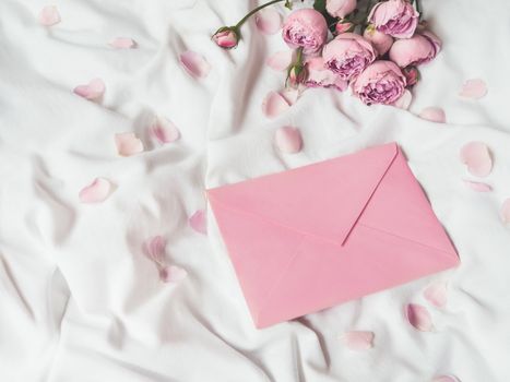 Roses and petals on crumpled white fabric. Natural elegant decoration. Romantic background with copy space on pink envelope. Flat lay.