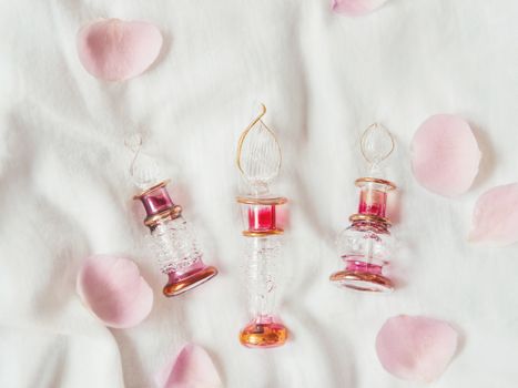 Three graceful bottles for perfume on white crumpled fabric. Top view on pink glass bottles with eastern ornament. Pink rose petals as decoration. Flat lay, still life.