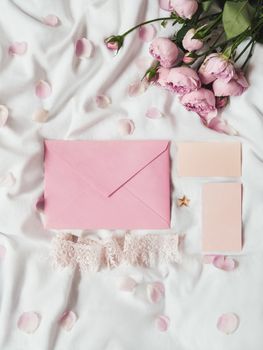 Roses and petals on crumpled white fabric. Natural elegant decoration. Romantic background with copy space on pink envelope and visit cards. Top view, flat lay.