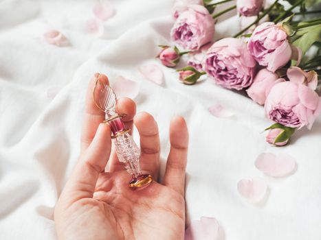 Graceful bottle for perfume or essential oil on white crumpled fabric. Woman holds pink glass bottle with eastern ornament. Pink rose bouquet and petals as decoration.